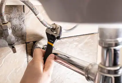 Plumber using an adjustable wrench to repair a sink supply line beneath a kitchen faucet.